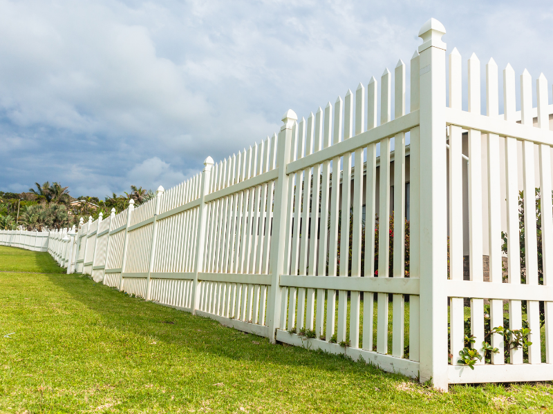 A white picket fence in front of a blue sky and green grass.