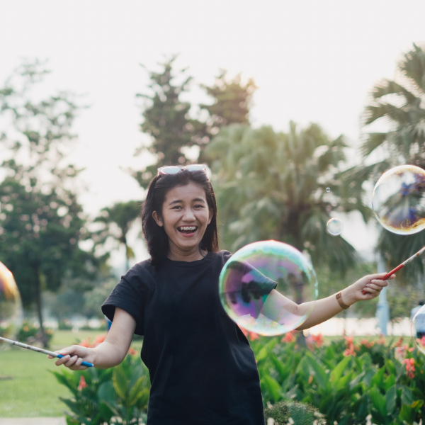 A woman smiles as she creates giant bubbles with a wand outdoors