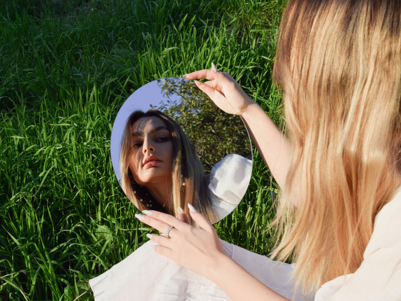 A woman looks into a mirror. She's sitting on a grass field.