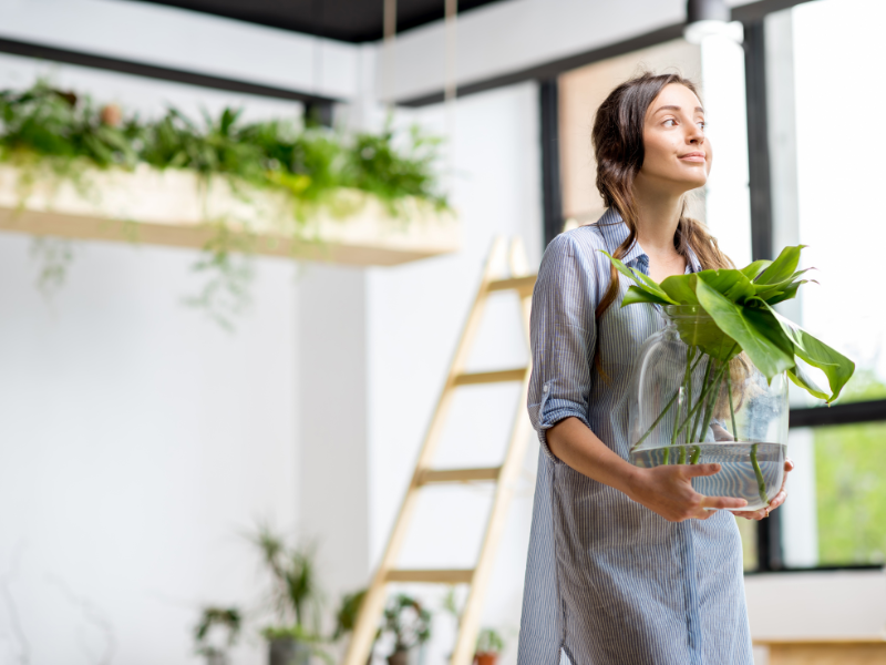 A woman smiles and looks proud as she holds a pot with a bright green plant in. She is surrounded by lush green plants.