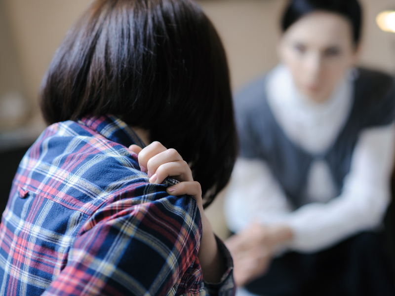 A woman is comforting herself, facing a therapist or supportive frined who is blurred into the background.