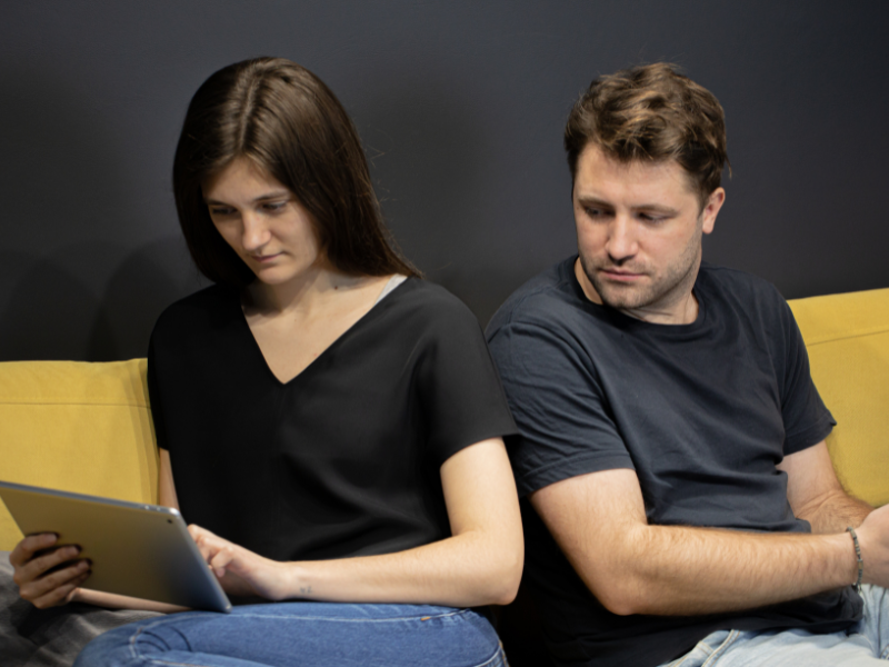 A woman is on her tablet, and her husband is watching what she is doing over her shoulder.