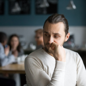 A man is standing alone in focus, looking anxious. A group of hii friends are blurred in the background.